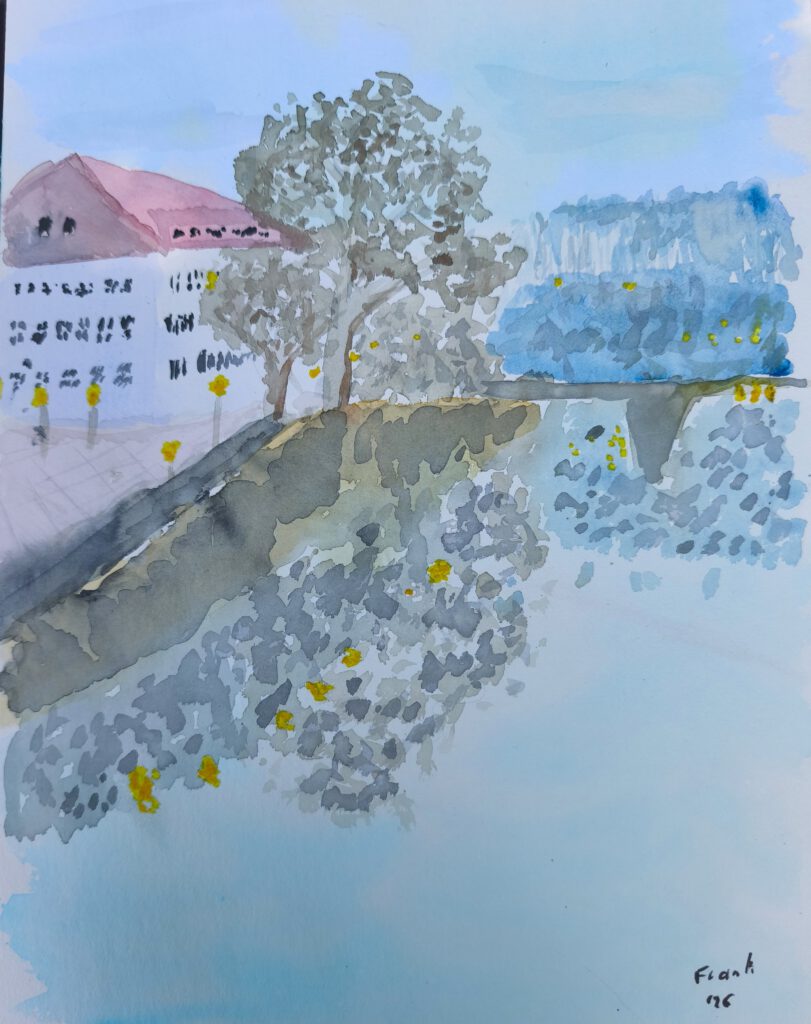 Ein großes helles Gebäude an einer Uferpromenade. Am Ufer steht ein großer Baun, der sich wie ein paar Laternen im Wasser spiegelt. in Hintergrund sieht man um eine Brücke herum weitere helle Punkte. Im Hintergurnd ein Wald. Für ein Bild mit Abendstimmung sind die Farben viel zu hell. 